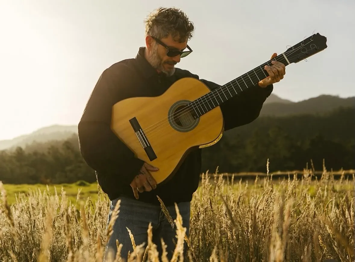 Juan Medina tocando la guitarra de pie en un campo, a contraluz de atardecer